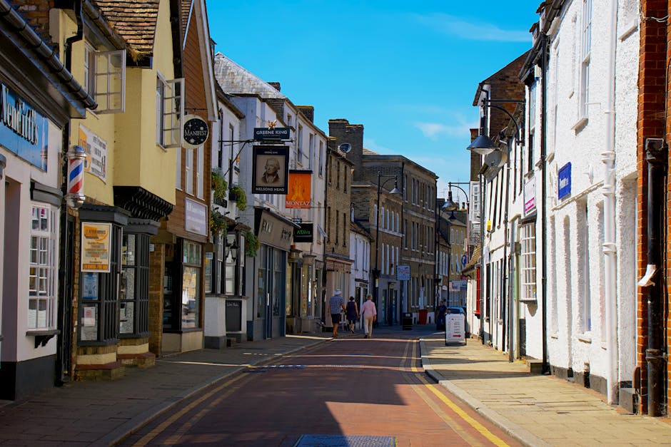 An empty narrow street in East Barnet with a clear blue sky overhead, showcasing a row of small retail shops and businesses housed in traditional brick and painted facades, with some storefronts featuring hanging signs, window displays, and planters. The street has a paved surface marked with double yellow lines along the curbs, and cast shadows from the buildings indicate bright sunlight. No actual moving or loading activity is visible, but the setting aligns with a typical location where house removals and furniture transport might take place, such as in preparation for customer moves or logistical planning by a professional removals service like Man with Van East Barnet. The image emphasizes the characteristic village scenery, compact street layout, and pedestrian-friendly environment typical of East Barnet Village, providing context for house relocation and packing processes in this community.