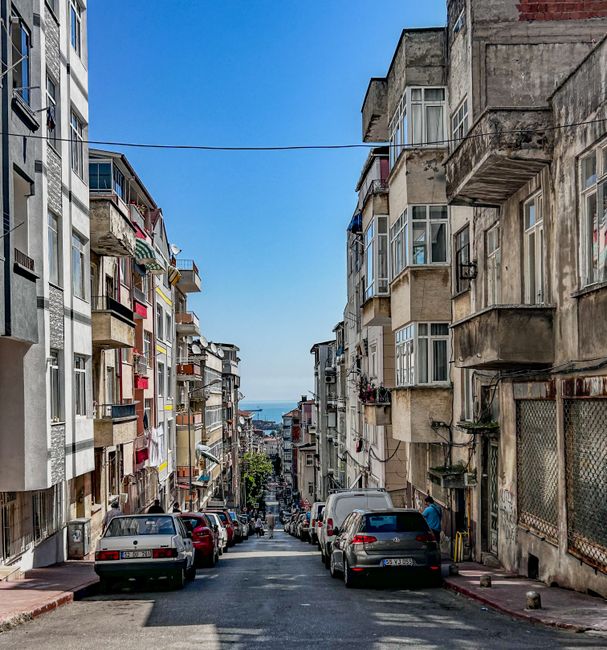 A narrow residential street during daytime with multi-storey apartment buildings on either side, featuring balconies with various plants and laundry hanging out. Several parked cars line both sides of the street, and a few individuals are visible near the buildings, possibly involved in a home relocation or moving process. The street slopes downward towards the sea, visible on the horizon under a clear blue sky. The buildings display a variety of materials, including concrete and brick, and some have weathered facades. This setting illustrates typical urban conditions for furniture transport and packing during house removals, with limited space for large vehicles like vans. Such environments require careful logistics, often facilitated by local moving services like Man with Van East Barnet, to ensure smooth loading and unloading of belongings in confined and narrow streets, supporting efficient home relocation and furniture transportation.