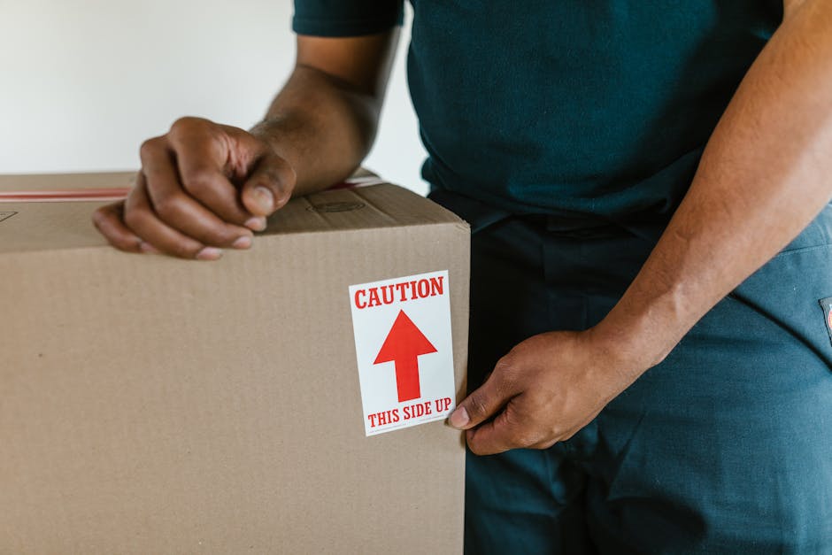 Close-up view of a person wearing a dark shirt and blue pants, handling a cardboard box during a house relocation process. The individual is gripping the box firmly while attaching a red and white caution sticker that reads 'CAUTION THIS SIDE UP' with an upward-pointing arrow, indicating proper orientation for transport. The box appears to be made of corrugated cardboard and is sealed at the top, ready for loading into a van or moving truck. The scene takes place indoors, with visible natural lighting and minimal background details, focusing on the action of careful handling and packing. This image exemplifies the packing and moving stages often managed by professional removals services such as Man with Van East Barnet, ensuring the safe transport of household items from an East Barnet property during home relocation or furniture transport activities.